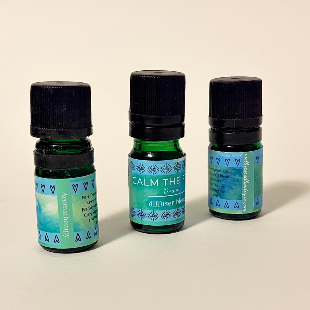 Three small glass essential oil diffuser bottles with black caps are arranged on a light beige background. The center bottle is upright and labeled 'CALM DOWN diffuser blend' with a turquoise label, while the two side bottles are turned, one shows the word aromatherapy and the other shows wellnessedboutique.com