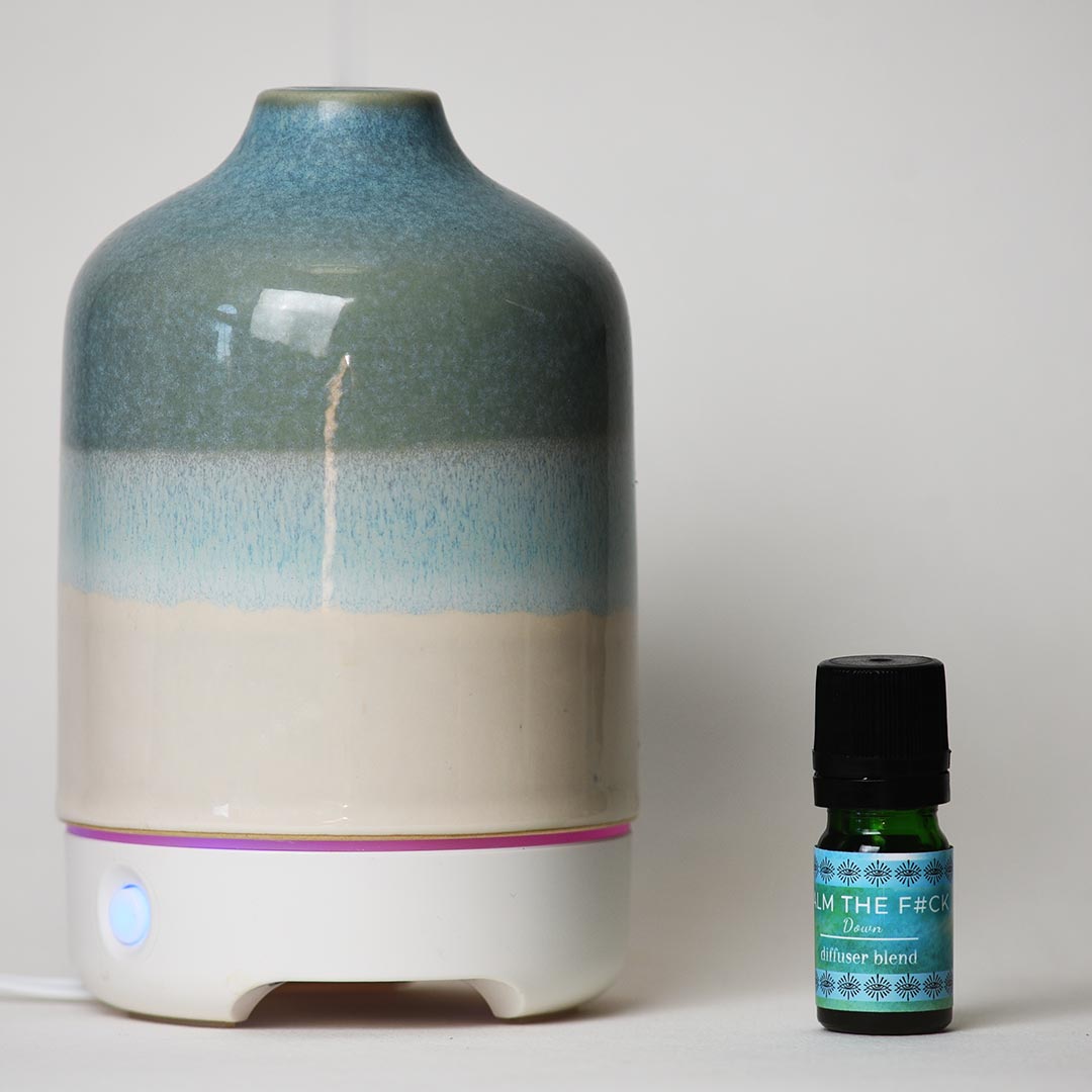 A ceramic diffuser with a blue, gray, and beige gradient glaze sits next to a small green essential oil bottle labeled 'Calm Down' Diffuser Blend on a light gray background.