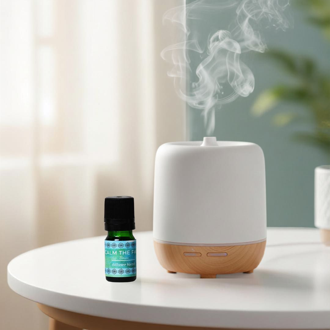 A white and light wood essential oil diffuser with misting steam, shown next to a small green bottle of Calm Down essential oil diffuser blend on a white table in a softly lit room.