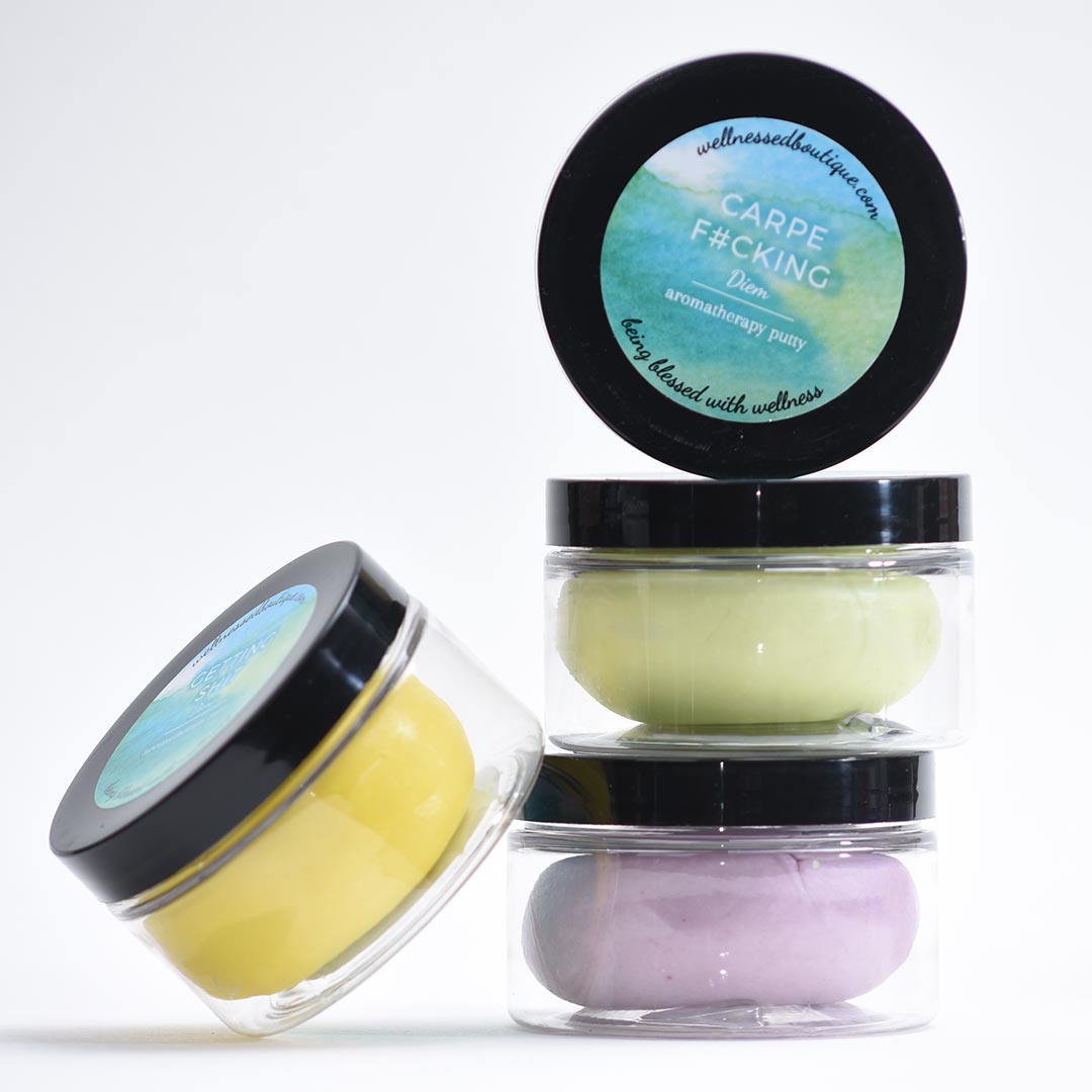 Four containers of Carpe Diem Aromatherapy Putty with black lids and colorful contents on a white background