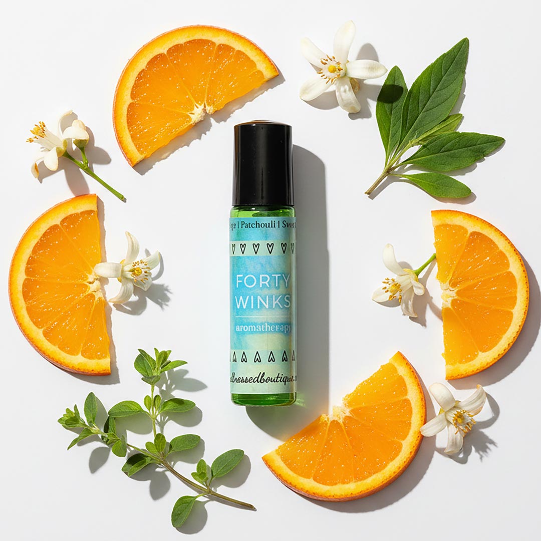 A bottle of Forty Winks sleep essential oil roll on surrounded by orange slices, sweet marjoram sprig, patchouli leaves, and orange blossoms
