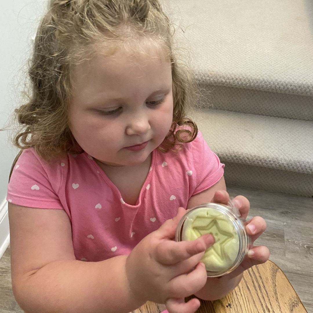 A young child with curly blonde hair wearing a pink shirt with small white hearts, holding a small clear jar containing Awesome Aromatherapy Putty. The child is seated indoors on a wooden chair near carpeted stairs.