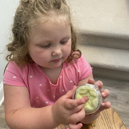 A young child with curly blonde hair wearing a pink shirt with small white hearts, holding a small clear jar containing Awesome Aromatherapy Putty. The child is seated indoors on a wooden chair near carpeted stairs.
